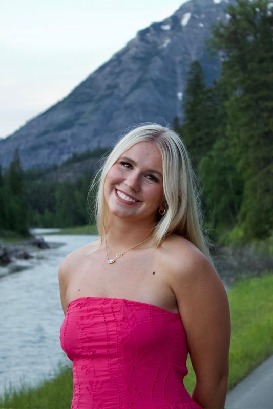 A young woman in a pink dress beside a river, a steep forested mountain rising behind her.