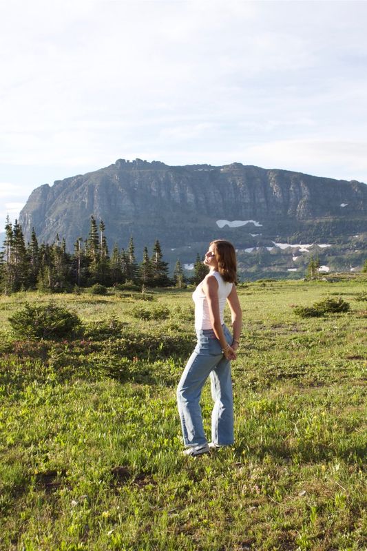 A young woman standing in an alpine meadow with her hands behind her back, looking up toward the light, a high cliff face behind her.