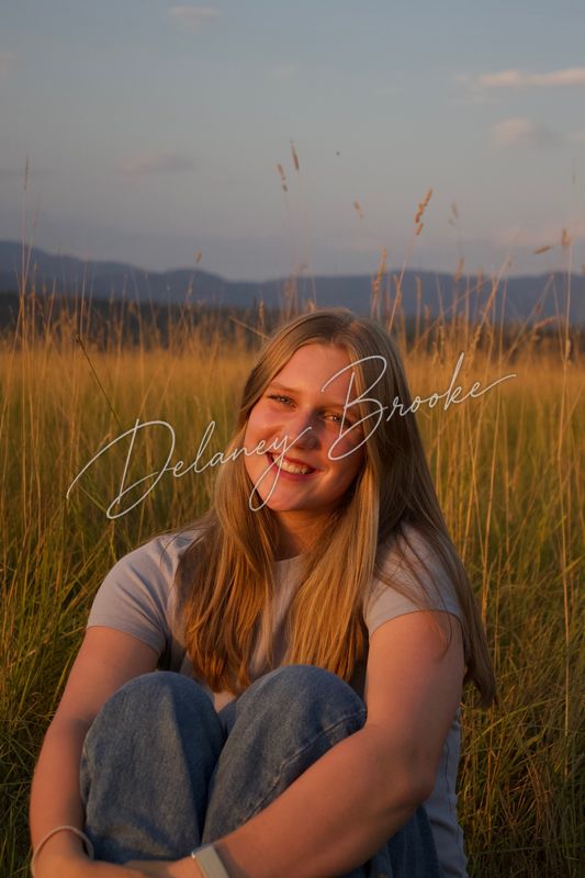 A young woman sitting in tall summer grass with her knees drawn up, smiling at the camera as the sun sets behind the mountains.