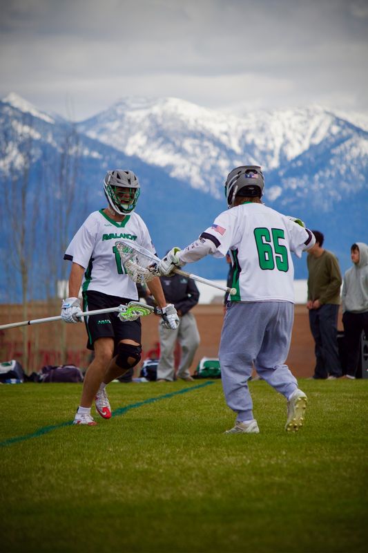 Two lacrosse players meeting on the field with snow-covered mountains behind them.