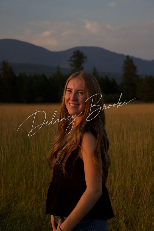A graduating senior standing in a summer field at dusk, turned over her shoulder toward the camera, a forested ridge behind her.