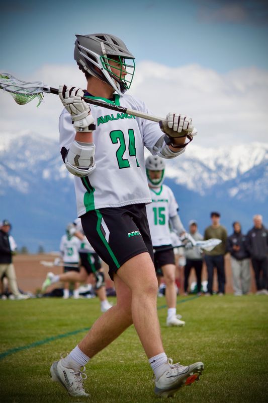 A lacrosse player carrying his stick across his shoulders, snow-covered peaks behind the field.