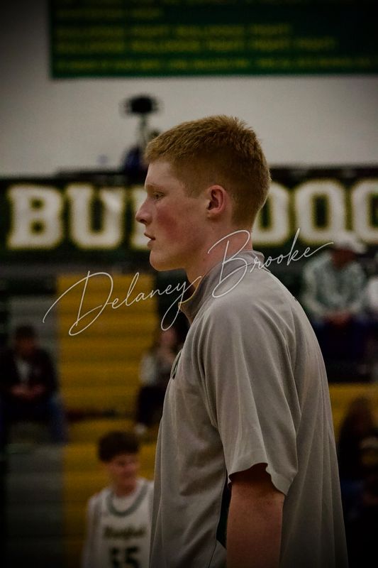 A player in a warm-up shirt watching from the sideline, the gym wall behind him.