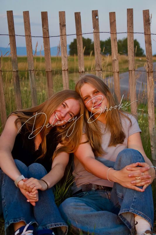 Two friends leaning their heads together against a weathered wooden fence at golden hour.