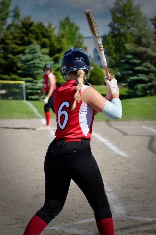 A softball batter seen from behind at the plate, bat raised, a runner leading off in the distance.