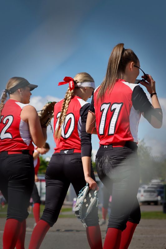 Three softball players standing together from behind in red jerseys, one lifting her sunglasses.