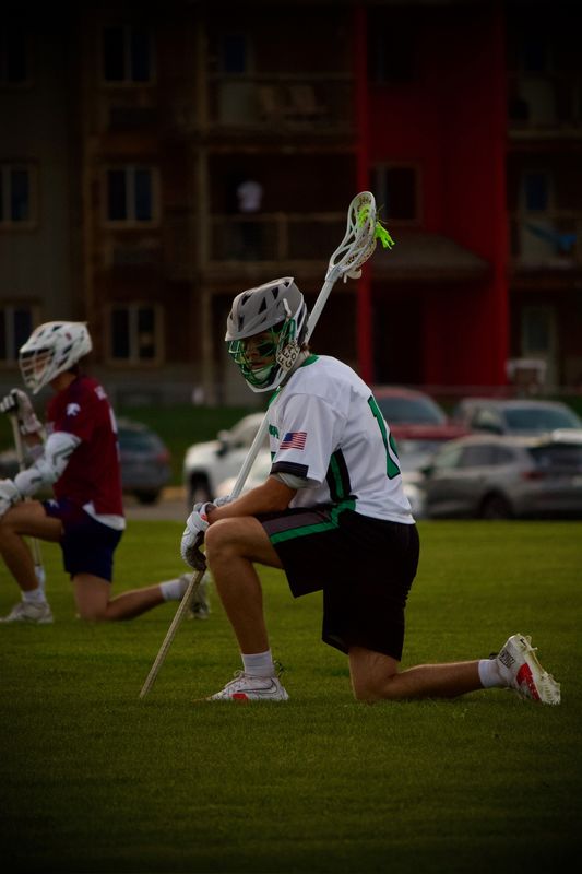 A lacrosse player taking a knee on the field, his stick planted upright beside him.