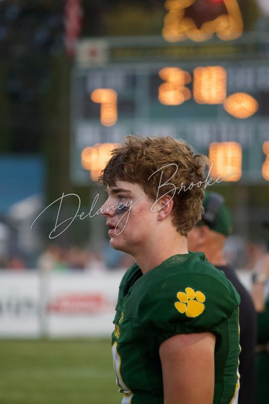 A football player in profile on the sideline at dusk, eye black under one eye, the lit scoreboard blurred behind him.
