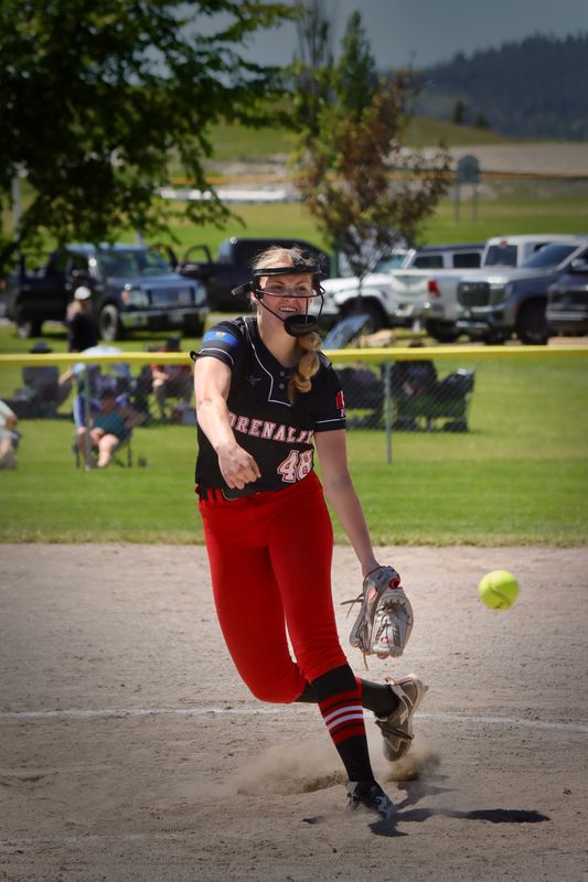 A softball pitcher mid-delivery, dust kicking up at her feet and the ball already in flight.