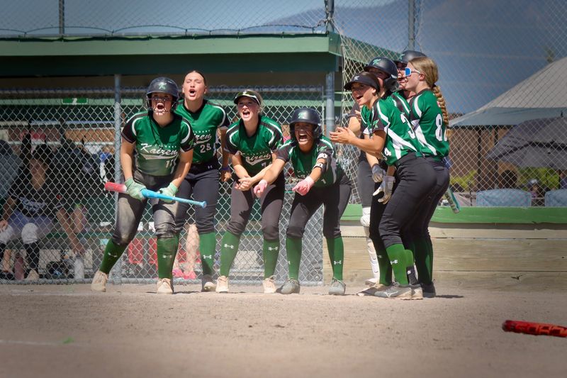 A softball team packed against the dugout fence mid-celebration, shouting toward the field.