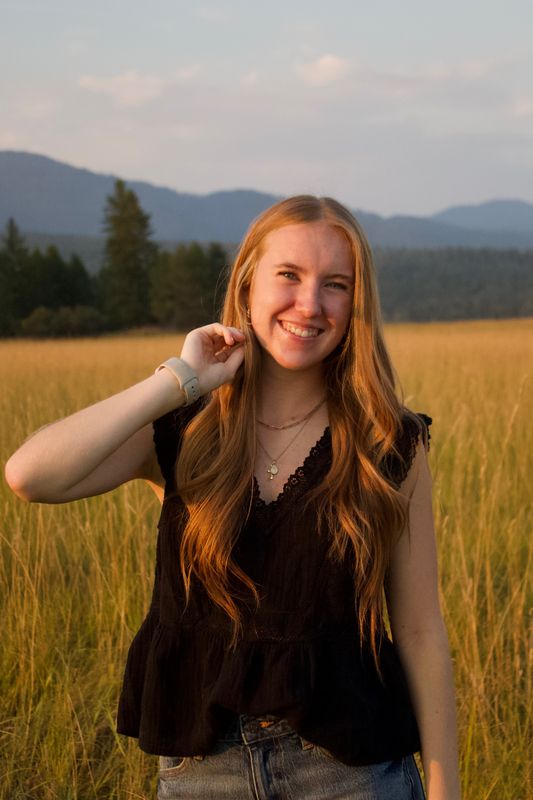 A graduating senior in a sunlit field at golden hour, mountains behind, smiling at the camera.