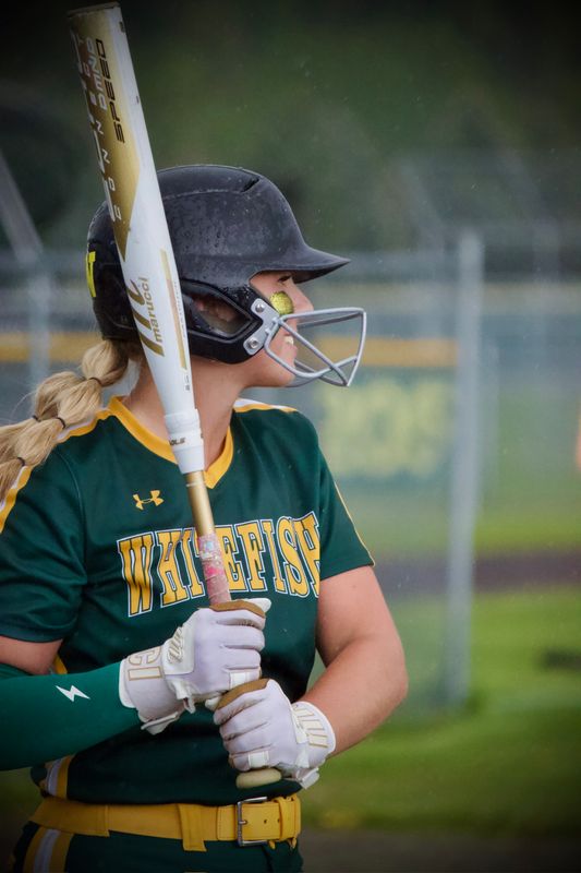 A softball batter waiting at the plate in the rain, bat raised, water beading on her helmet.